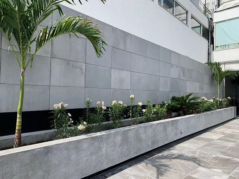 Jardinera de concreto aparente con palmas y plantas en flor frente a un muro de paneles de concreto, en el exterior del Club Alpino Chipinque