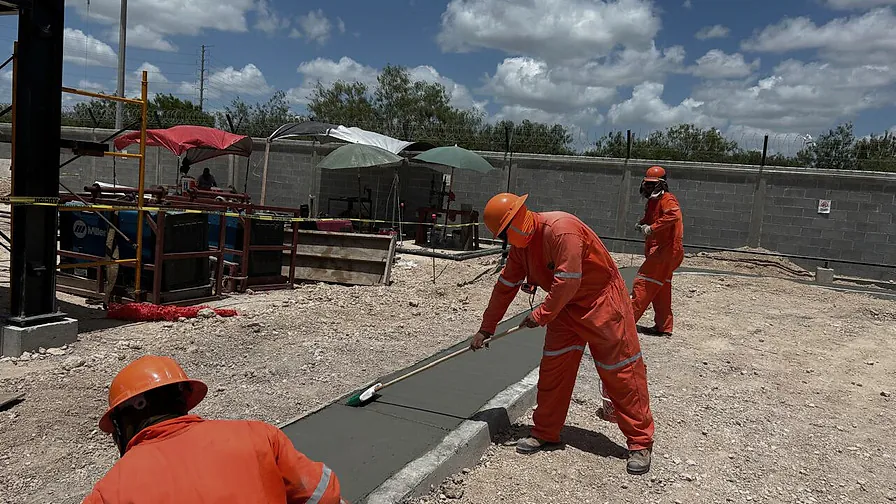 Trabajadores de overol naranja dan acabado escobillado a una banqueta recién colada en la Terminal K19