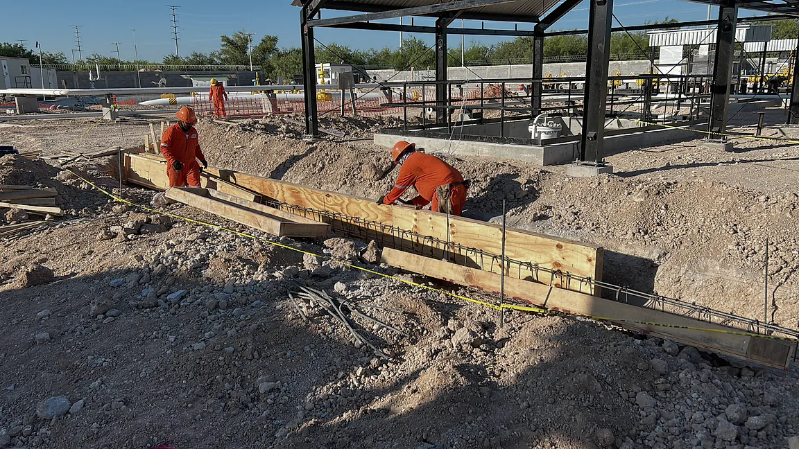 Dos trabajadores de overol naranja arman la cimbra de madera y el acero de refuerzo a nivel de terreno en la Terminal K19